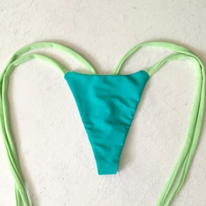 May include: A turquoise bikini bottom with lime green ties. The triangle-shaped bottom is made of a smooth fabric. The long ties extend from the sides and are knotted at the ends. The background is a textured white surface.