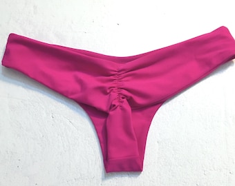 May include: A pair of hot pink bikini bottoms. The bottoms are ruched in the front and have a low-rise cut. The fabric is smooth and appears to be a solid colour. The background is a plain white wall.
