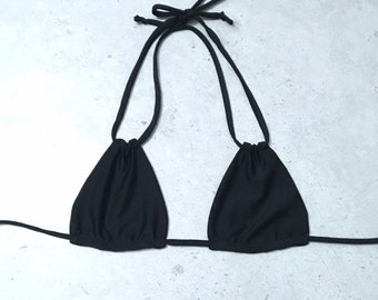 Puede incluir: Un top de bikini negro liso con dos copas triangulares con tela fruncida en los bordes superiores. La prenda incluye finos tirantes negros que se extienden desde la parte superior de cada copa para atarse al cuello y una tira larga en la base.