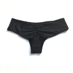 K&ouml;nnte beinhalten: Eine einfarbige schwarze Bikini-Unterhose mit einer gerafften, gef&auml;ltelten Mittelnaht am R&uuml;ckenteil. Das Kleidungsst&uuml;ck hat einen breiten, glatten Bund und einen tief sitzenden Schnitt, gefertigt aus einem schlichten, matten Stoff ohne Muster.