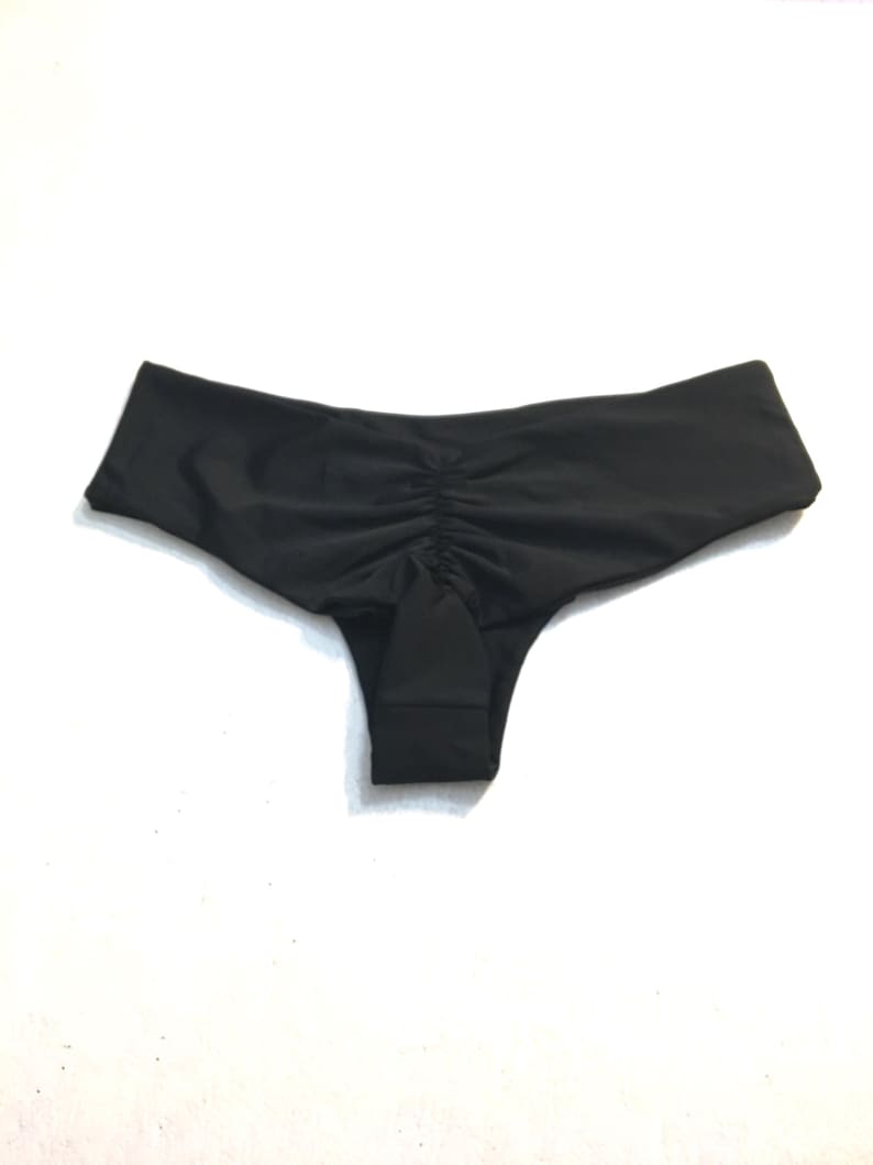 K&ouml;nnte beinhalten: Eine einfarbige schwarze Bikini-Unterhose mit einer gerafften, gef&auml;ltelten Mittelnaht am R&uuml;ckenteil. Das Kleidungsst&uuml;ck hat einen breiten, glatten Bund und einen tief sitzenden Schnitt, gefertigt aus einem schlichten, matten Stoff ohne Muster.