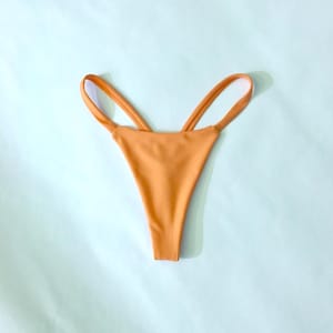 Puede incluir: Una braga de bikini de color naranja s&oacute;lido con un dise&ntilde;o de pierna alta y detalles de tiras dobles finas en los laterales. La prenda tiene un acabado liso y mate, con una construcci&oacute;n minimalista que muestra una est&eacute;tica limpia y sencilla.
