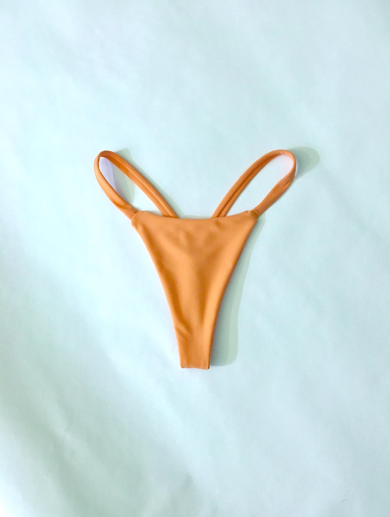Puede incluir: Una braga de bikini de color naranja s&oacute;lido con un dise&ntilde;o de pierna alta y detalles de tiras dobles finas en los laterales. La prenda tiene un acabado liso y mate, con una construcci&oacute;n minimalista que muestra una est&eacute;tica limpia y sencilla.