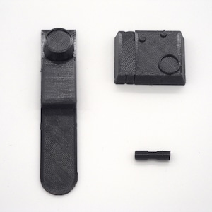 Puede incluir: Tres piezas negras impresas en 3D para un kit de maquetas. Las piezas incluyen una pieza larga, delgada y rectangular con una perilla redonda en la parte superior, una pieza rectangular más pequeña con un agujero redondo y dos pequeñas protuberancias, y una pieza corta, delgada y cilíndrica.