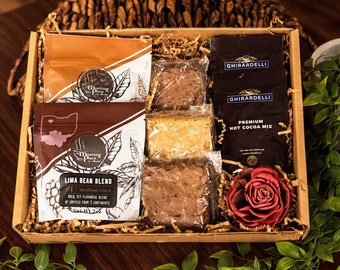 May include: A brown gift box filled with a coffee blend, hot cocoa mix, brownies, and a red rose. The box is filled with brown packing paper and has a brown lid. The coffee blend is in a brown bag with the words "The Meeting Place" and "Lima Bean Blend" printed on it. The hot cocoa mix is in a black bag with the words "Ghirardelli" and "Premium Hot Cocoa Mix" printed on it. The brownies are in clear plastic bags. The rose is red and has a green stem.