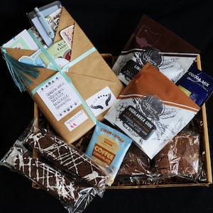 May include: A gift basket filled with various treats and items. It includes packaged brownies with white icing, coffee and cocoa mix packets, and a wrapped package with a teal ribbon. The package has text that reads "Guardian Icons, Abscaliptic, Serenity, Resilience, Strong Fine."