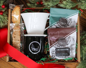 May include: A gift box containing a black coffee mug with the text "Meeting Place", a white coffee dripper, two bags of coffee, and biscotti. The box is decorated with a red ribbon and Christmas greenery.