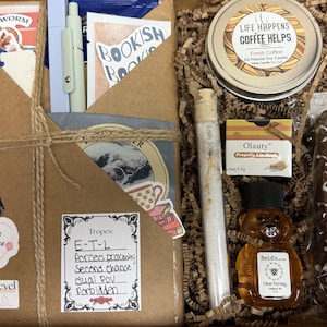 May include: A gift box filled with various items, including a candle, honey, bath salts, a cookie, and a book. The box is decorated with twine and a blue tassel. The box includes a tag that says "Genre Romance" and "Spice Level".