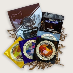 K&ouml;nnte beinhalten: Eine Sammlung von Tee- und Kaffees&auml;ckchen in verschiedenen Geschmacksrichtungen, darunter Earl Grey, English Breakfast, Brombeer-Salbei, Kamille-Zitrone und Limabohnen-Mischung. Die S&auml;ckchen sind auf einem Bett aus braunem, zerknittertem Papier angeordnet.