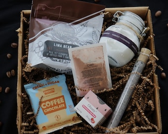 May include: A gift box filled with coffee, bath products, and lip balm. The box contains a bag of Lima Bean coffee, a jar labeled "Harvest + Harmony", a packet of "Calm" powder, a tube, a packet of "Coffee Chocolate" and a box of "Olauty" lip balm.