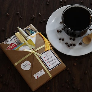 May include: A cup of black coffee on a white saucer with a wooden spoon, next to a brown paper package tied with a yellow ribbon. The package contains various items, including stickers and cards. Coffee beans are scattered around.
