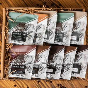 May include: A gift box filled with various coffee bean blends. The coffee bags have different colors and labels, with text such as "Jamaican Me Crazy" and "Lima Bean Blend". The box sits on a wooden surface with scattered coffee beans.