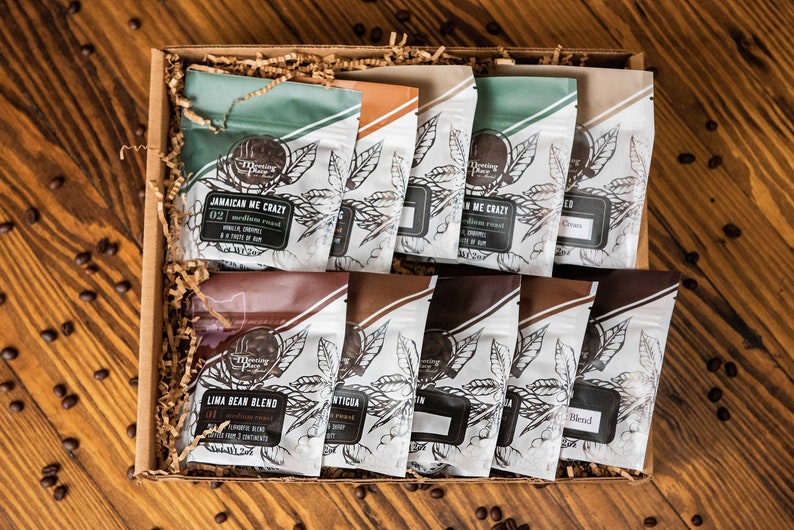 May include: A gift box filled with various coffee bean blends. The coffee bags have different colors and labels, with text such as "Jamaican Me Crazy" and "Lima Bean Blend". The box sits on a wooden surface with scattered coffee beans.
