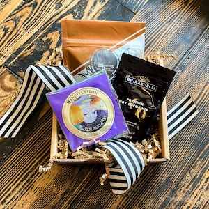 K&ouml;nnte beinhalten: Eine Geschenkbox gef&uuml;llt mit braunen Papierschnitzeln und einem schwarz-wei&szlig; gestreiften Band. Die Box enth&auml;lt drei Artikel: eine violette Teebeutelverpackung mit dem Text "Mango Ceylon The Republic of Tea Round Black Tea Bag", eine braune Verpackung mit dem Text "Meeting Place" und eine schwarze Verpackung mit dem Text "Ghirardelli Premium Hot Cocoa Mix".