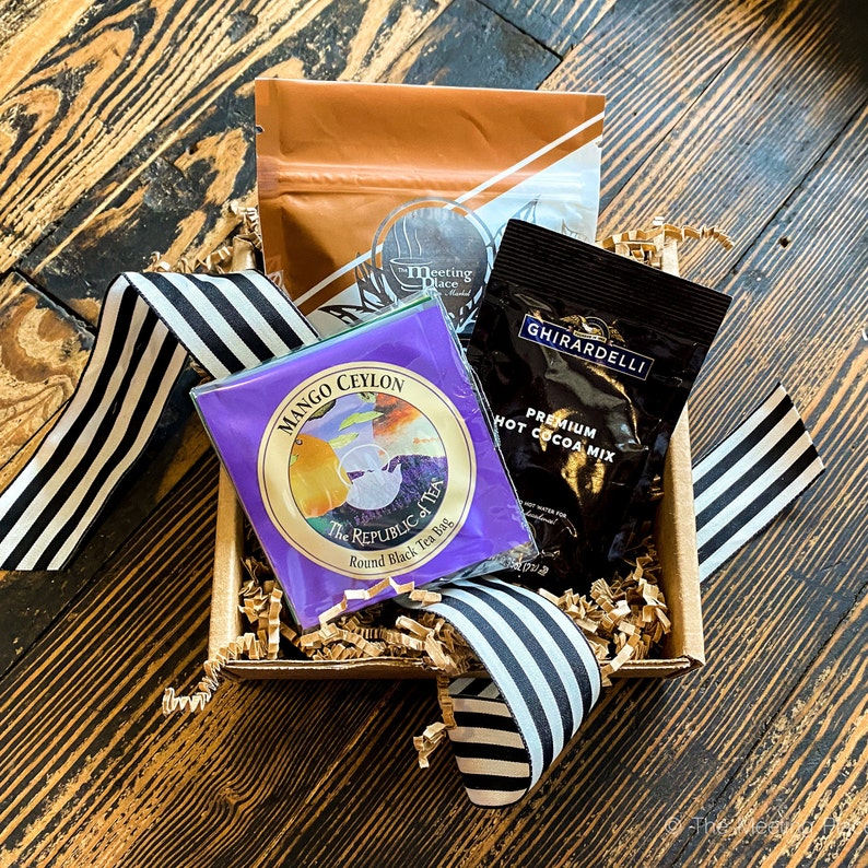 K&ouml;nnte beinhalten: Eine Geschenkbox gef&uuml;llt mit braunen Papierschnitzeln und einem schwarz-wei&szlig; gestreiften Band. Die Box enth&auml;lt drei Artikel: eine violette Teebeutelverpackung mit dem Text "Mango Ceylon The Republic of Tea Round Black Tea Bag", eine braune Verpackung mit dem Text "Meeting Place" und eine schwarze Verpackung mit dem Text "Ghirardelli Premium Hot Cocoa Mix".