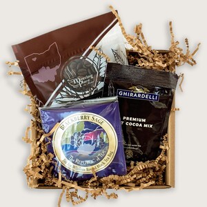 K&ouml;nnte beinhalten: Eine braune Geschenkbox, gef&uuml;llt mit braunen Papierschnitzeln, enth&auml;lt einen braunen Teebeutel mit dem Text "Blackberry Sage" und den Worten "The Republic of Tea" in einem Kreis, ein braunes P&auml;ckchen mit hei&szlig;er Schokolade mit dem Text "Ghirardelli Premium Hot Cocoa Mix" und einen braunen Teebeutel mit dem Text "The Meeting Place" in einem Kreis.