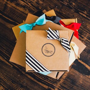 K&ouml;nnte beinhalten: Ein Stapel brauner Geschenkboxen mit blauen, schwarzen und roten Schleifen. Die oberste Box hat ein schwarz-wei&szlig; gestreiftes Band und ein Logo mit der Aufschrift "The Meeting Place in Madrid".