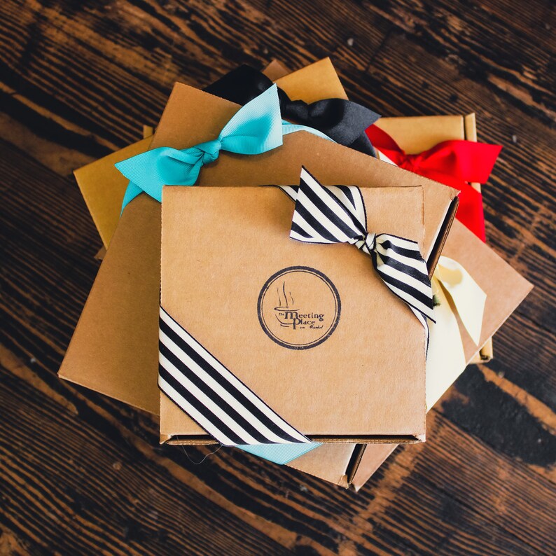 K&ouml;nnte beinhalten: Ein Stapel brauner Geschenkboxen mit blauen, schwarzen und roten Schleifen. Die oberste Box hat ein schwarz-wei&szlig; gestreiftes Band und ein Logo mit der Aufschrift "The Meeting Place in Madrid".