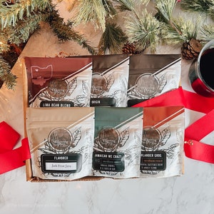 May include: A gift box of six coffee bean bags with various flavors, including "Lima Bean Blend" and "Jamaican Me Crazy." The bags are in a cardboard box, with a red ribbon and pine branches in the background. A glass of red liquid is also visible.