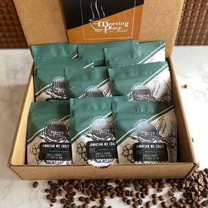 May include: A brown cardboard box filled with 12 green pouches of coffee. The pouches are labeled "Jamaican Me Crazy" with the text "02 medium roast" and "Vanilla, Caramel & a Taste of Rum". The box is open and there are coffee beans scattered on a white surface.