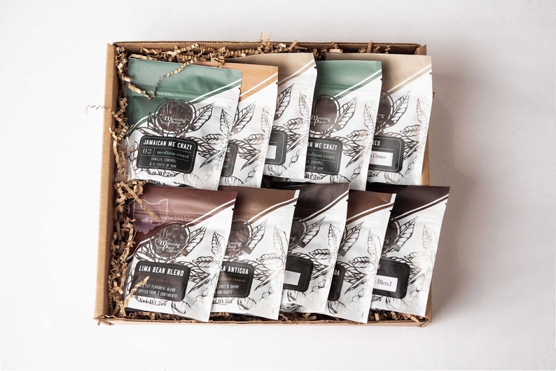 May include: A gift box filled with various coffee bean bags. The bags have a white background with a leaf design and text. The box is brown and filled with shredded paper. The coffee bags have different colors and text labels.