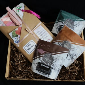 May include: A gift box with a brown paper wrap, pink ribbon, and a pink tassel. Inside are pink pens, a palette, and floral stickers. Coffee bags with the text "Lima Bean" and "Meeting Place" are also in the box.