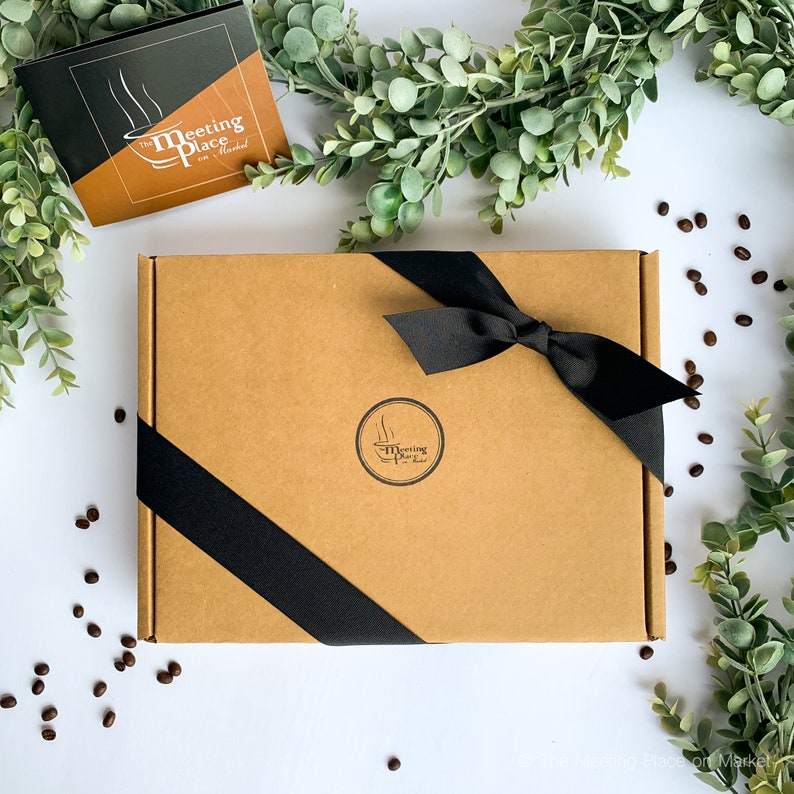 May include: A brown cardboard gift box with a black ribbon and bow. The box has a circular logo that says "The Meeting Place on Market". A card with the same logo is on the left. Coffee beans and greenery surround the box.