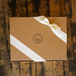 May include: A rectangular, brown cardboard box tied with a cream-colored satin ribbon. The ribbon is knotted on top and wraps diagonally across the box. A circular logo with the text "Meeting Place" is printed on the box.