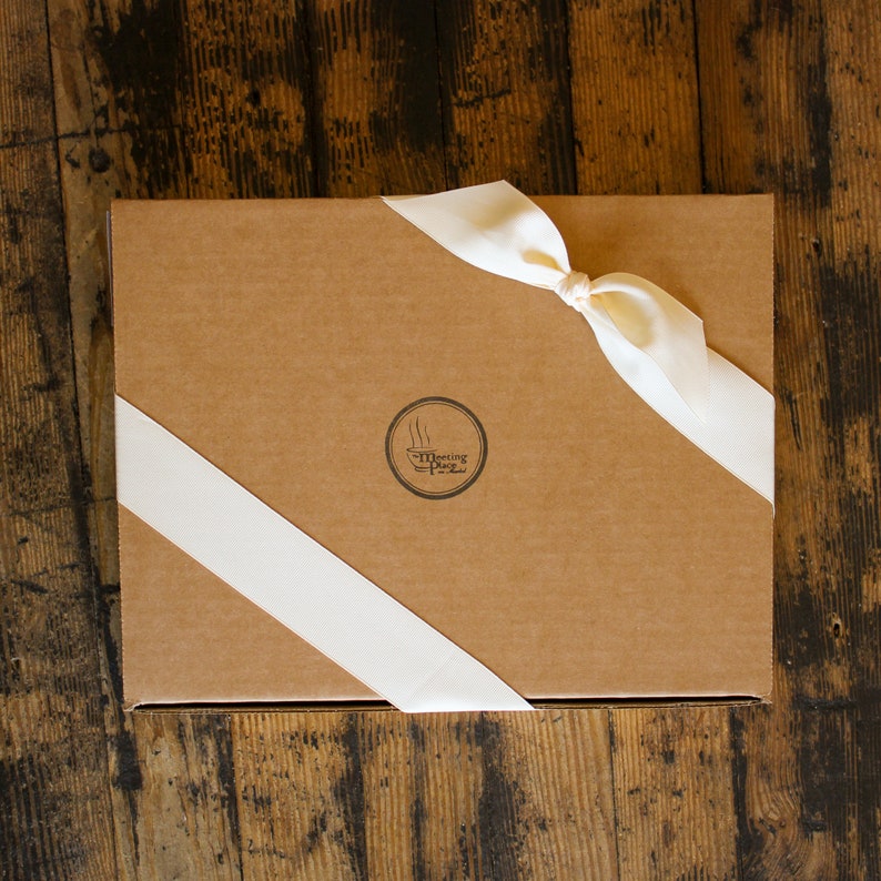 May include: A rectangular, brown cardboard box tied with a cream-colored satin ribbon. The ribbon is knotted on top and wraps diagonally across the box. A circular logo with the text "Meeting Place" is printed on the box.