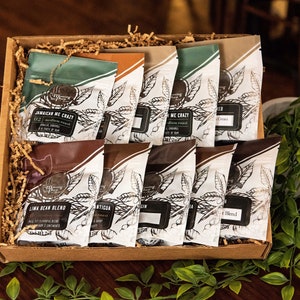 May include: A gift box filled with various coffee bean blends. The coffee bags have a white background with a black leaf design and text. The box is brown and filled with brown paper filler. The coffee bags have different colored tops, including green, brown, and tan.