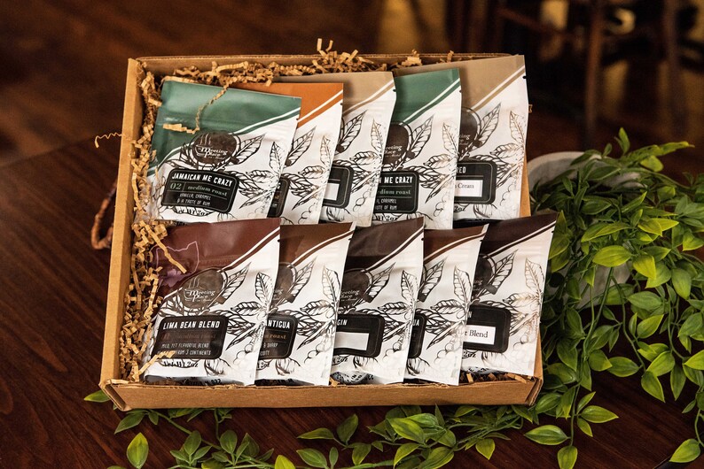 May include: A gift box filled with various coffee bean blends. The coffee bags have a white background with a black leaf design and text. The box is brown and filled with brown paper filler. The coffee bags have different colored tops, including green, brown, and tan.