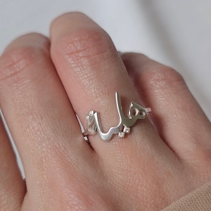 May include: A silver ring featuring a delicate Arabic script design with small clear stones.