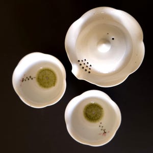 May include: A white ceramic tea set consisting of one teapot and two small cups. Each piece features a scalloped edge and a delicate interior design of a green circular pattern with a small branch of pink flowers and dark buds painted on the side.