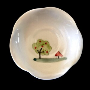 May include: A small, white, scalloped-edge ceramic bowl featuring a hand-painted illustration at the centre. The design depicts a green tree with red fruit, a small red-roofed structure, and a horizontal green line representing the ground.