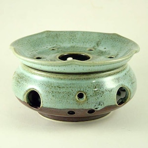 May include: A teal and brown ceramic oil warmer. The two-piece design features a top dish with multiple holes and a curved edge. The base has several circular openings. The warmer has a speckled finish and a brown base.
