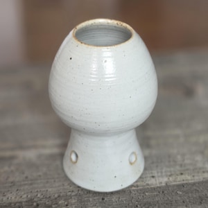 May include: A speckled off-white ceramic oil burner with a bulbous top section and a flared base. The piece features a circular opening at the top and two small, circular ventilation holes positioned symmetrically near the bottom of the base.