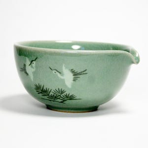 May include: A light green ceramic tea pitcher with a crackle glaze finish. The exterior features a hand-painted design of two white cranes in flight above dark green pine branches. The vessel has a small, integrated pouring spout on the rim.