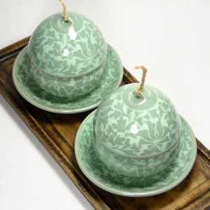 May include: Two matching green ceramic lidded bowls with floral patterns, each resting on a saucer. The bowls have a spherical shape and a small string attached to the top. The set is displayed on a wooden tray.
