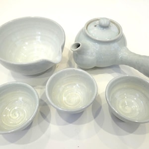 May include: A light gray ceramic tea set, including a teapot with a long handle, a serving bowl with a spout, and three small teacups. The set has a minimalist design and a smooth, glazed finish, perfect for serving tea.