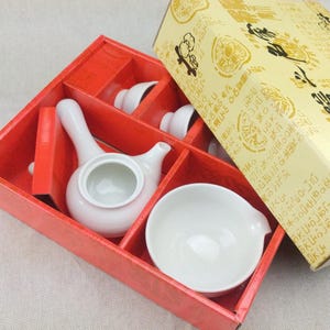 May include: A white ceramic teapot with a side handle and a matching white bowl are nestled inside a red cardboard box. A light yellow lid with black printed text and graphic illustrations rests partially over the open box container.