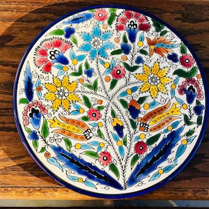 May include: A round, hand-painted ceramic plate with a vibrant floral design. The plate features a white base with a blue rim and is adorned with colorful flowers in shades of red, yellow, blue, and orange. Green leaves and stems complete the intricate pattern.