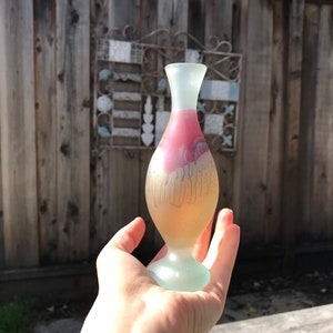 May include: A frosted glass vase with a flared top and a rounded base. The vase features a gradient of colors, with a pink and white interior and a pale yellow exterior. The vase is held in a hand against a wooden fence background.