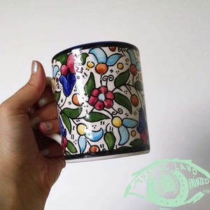 May include: A ceramic mug with a floral design. The mug features a white base with a pattern of colorful flowers, green leaves, and orange accents. The rim is dark blue. The text "MYSTERY LAND PAINTED OWN & ADORE" is visible.