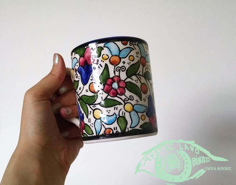 May include: A ceramic mug with a floral design. The mug features a white base with a pattern of colorful flowers, green leaves, and orange accents. The rim is dark blue. The text "MYSTERY LAND PAINTED OWN & ADORE" is visible.