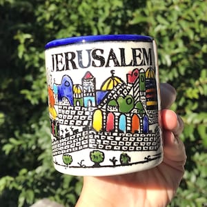 Pode incluir: Uma caneca de cerâmica com uma borda azul e a palavra "JERUSALEM" em letras pretas. A caneca apresenta uma ilustração colorida da arquitetura de Jerusalém, incluindo edifícios com detalhes azuis, amarelos, vermelhos e verdes. A caneca é segurada em uma mão.