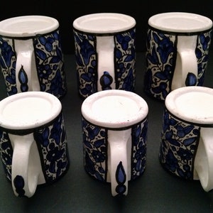 May include: Six ceramic mugs with white handles and rims. The mugs are decorated with a blue floral pattern on a white background. The mugs are arranged on a dark surface.