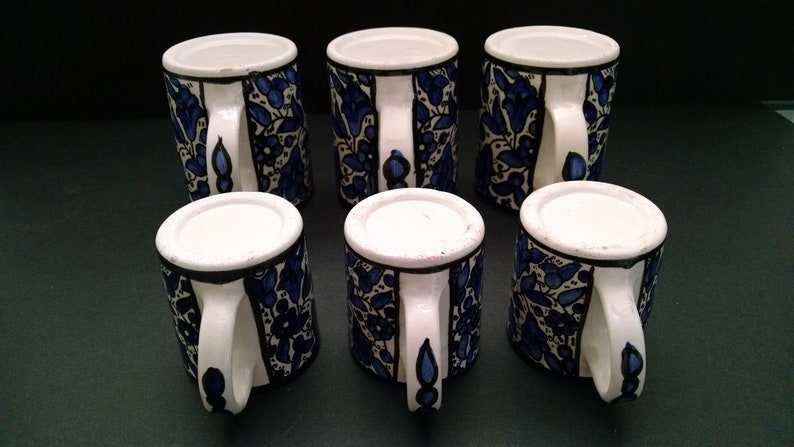 May include: Six ceramic mugs with white handles and rims. The mugs are decorated with a blue floral pattern on a white background. The mugs are arranged on a dark surface.