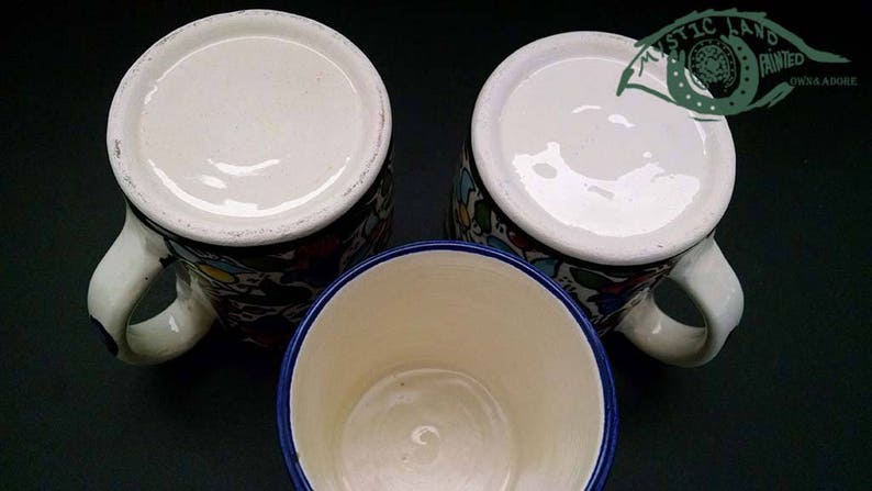May include: Three ceramic mugs with white interiors and handles. The mugs have colorful floral patterns and a blue rim. The image includes the text "MYSTIC LAND PAINTED OWNA ADOBE".