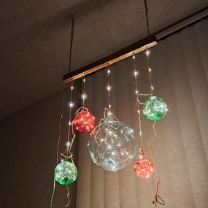May include: A decorative hanging light fixture with six glass orbs in red, green, and clear, each lit with small white lights. The orbs are suspended from a wooden beam by chains, creating a festive and warm atmosphere.