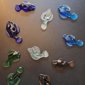 May include: A collection of glass fish figurines in various colors, including blue, green, and clear. The fish have detailed features and are arranged on a dark surface. The figurines are small, decorative items.